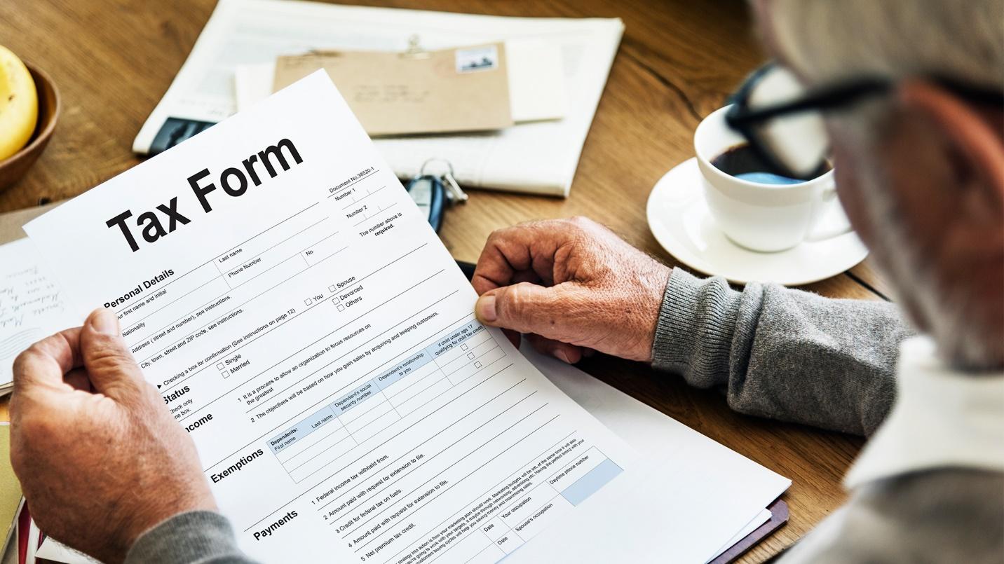 An elderly man studies a tax form at a table beside letters and a cup of coffee in a section about how to apply for tax breaks. 