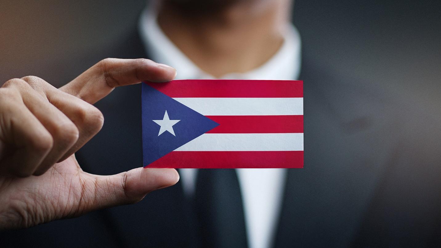 A man in a suit holds a business card that depicts the flag of Puerto Rico in a section about the low taxes and other advantages that come with moving to Puerto Rico.