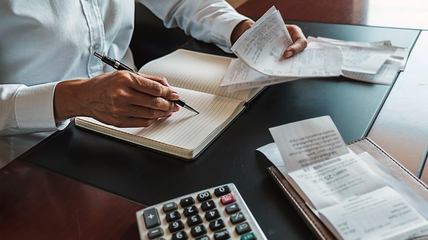 Person writing in a notebook at a desk while holding receipts.