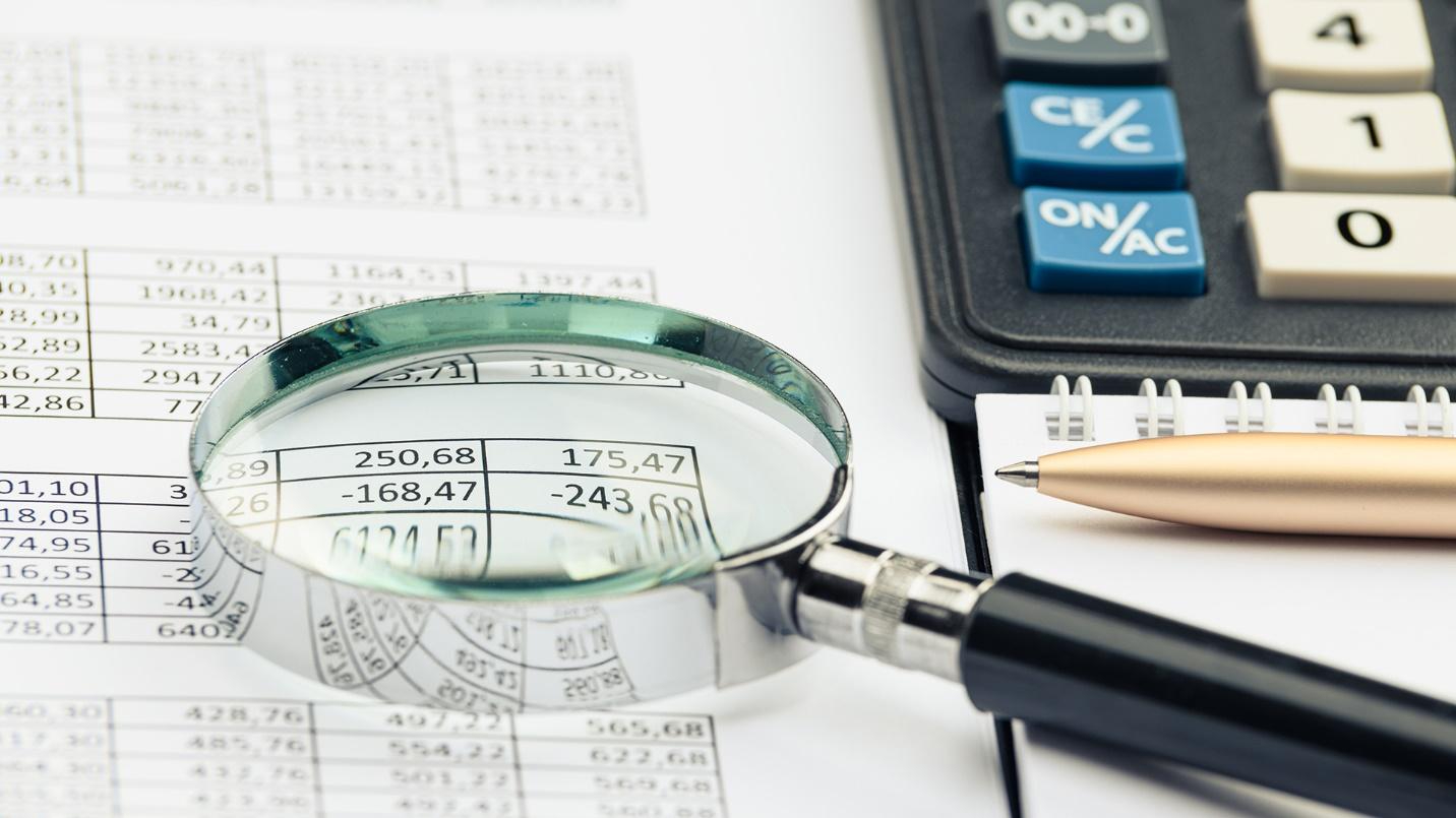 A magnifying glass resting on a sheet with tables of numbers, next to a calculator and pen.