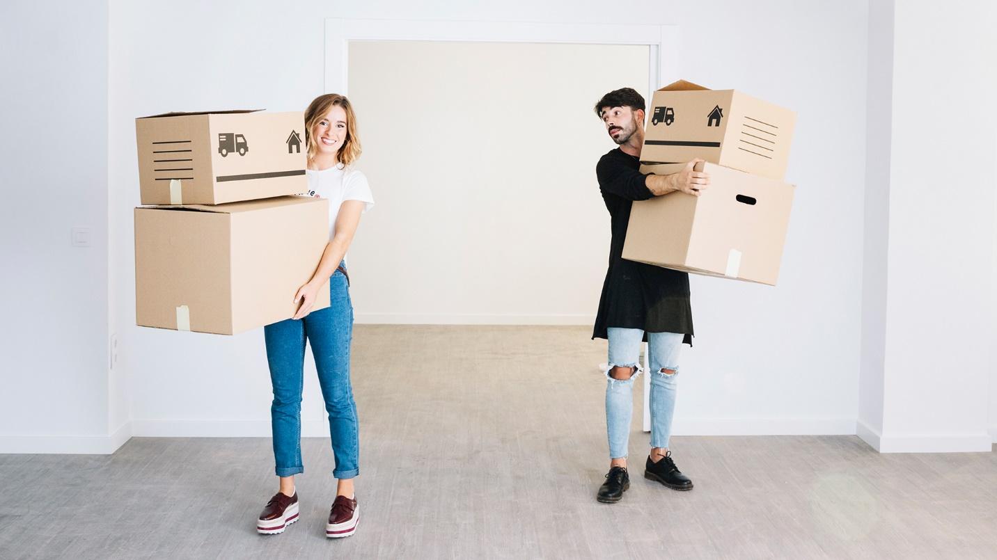 A man and woman carry cardboard boxes into an empty room in a section about whether Americans need passports to relocate to Puerto Rico.
