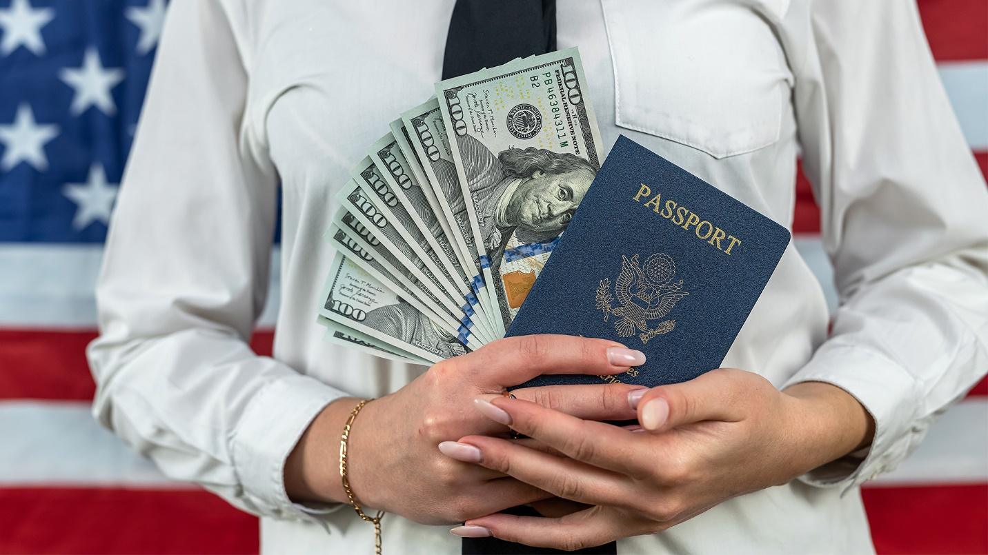 A woman wearing a white blouse holds a passport and several hundred-dollar bills while standing in front of an American flag in a section describing the fees for passport applications.

