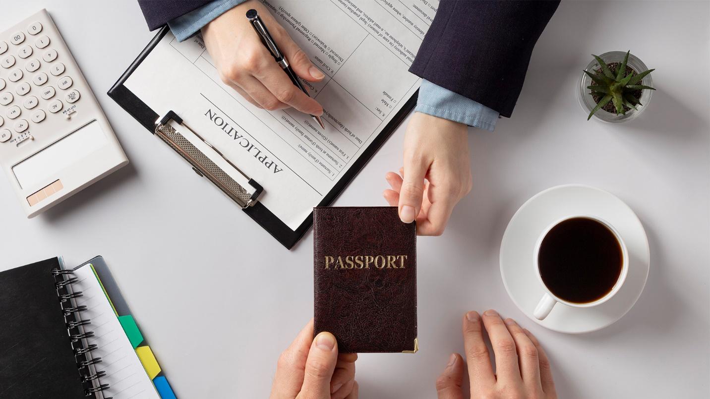 One person hands a passport to another over a table that holds paperwork, a calculator, coffee, and a plant in a section about passport offices in Puerto Rico.
