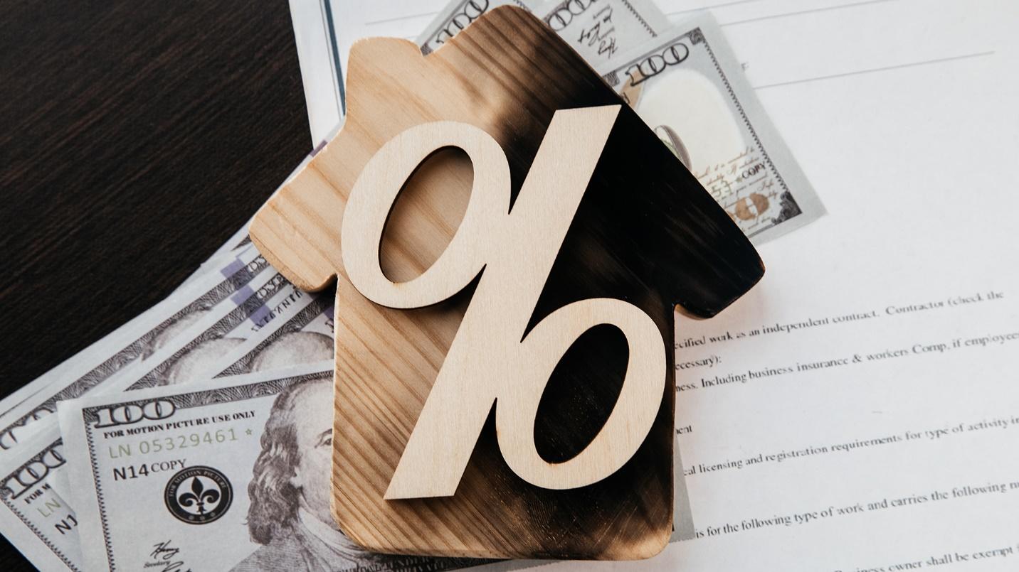 A percent sign on a house-shaped wooden board surrounded by $100 bills and paperwork in a section about capital gains tax savings for bona fide residents of Puerto Rico.