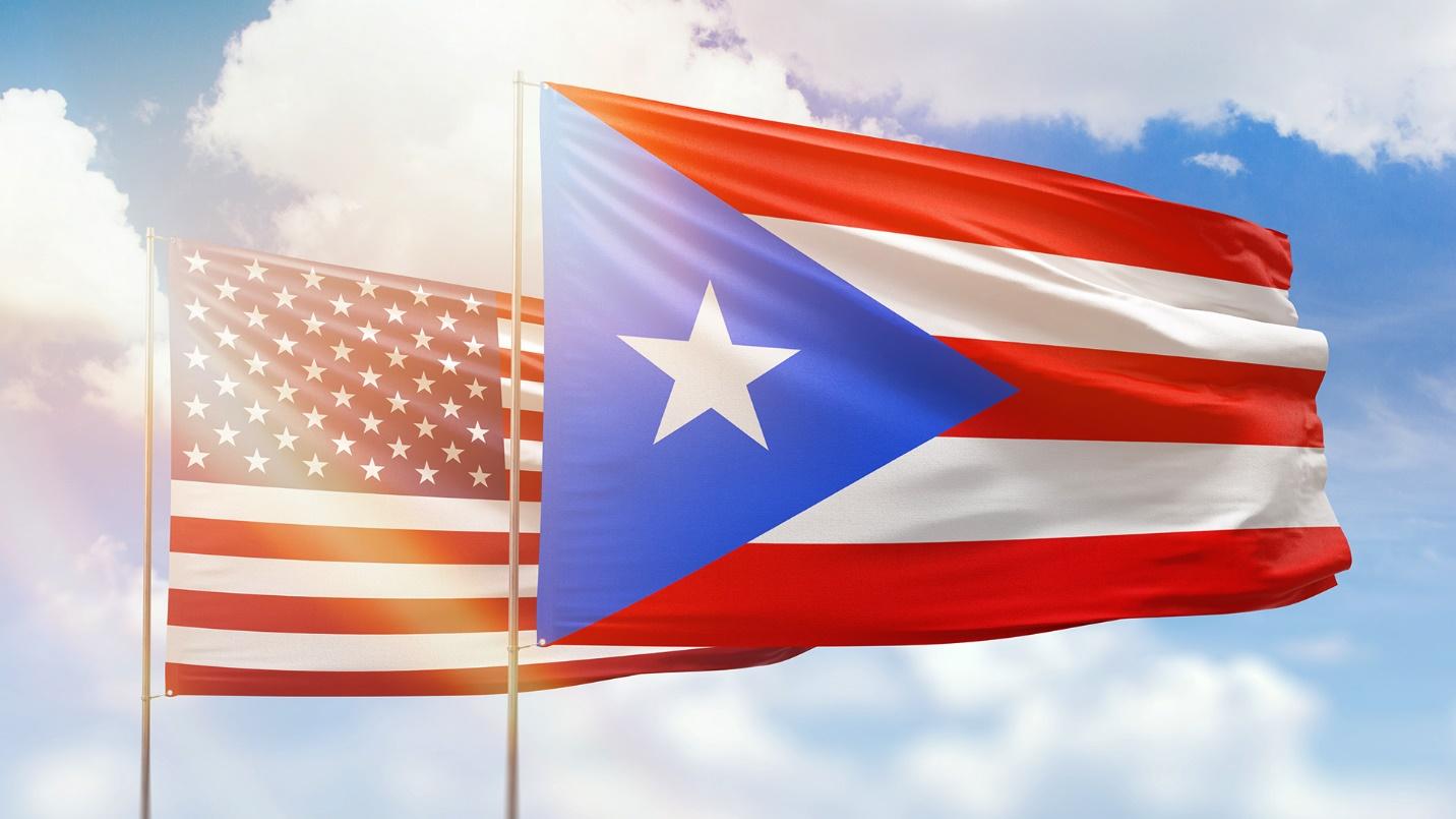 The U.S. and Puerto Rican flags illuminated by sunlight against a cloudy sky background in a section about how the United States and Puerto Rico treat taxes on cryptocurrency.