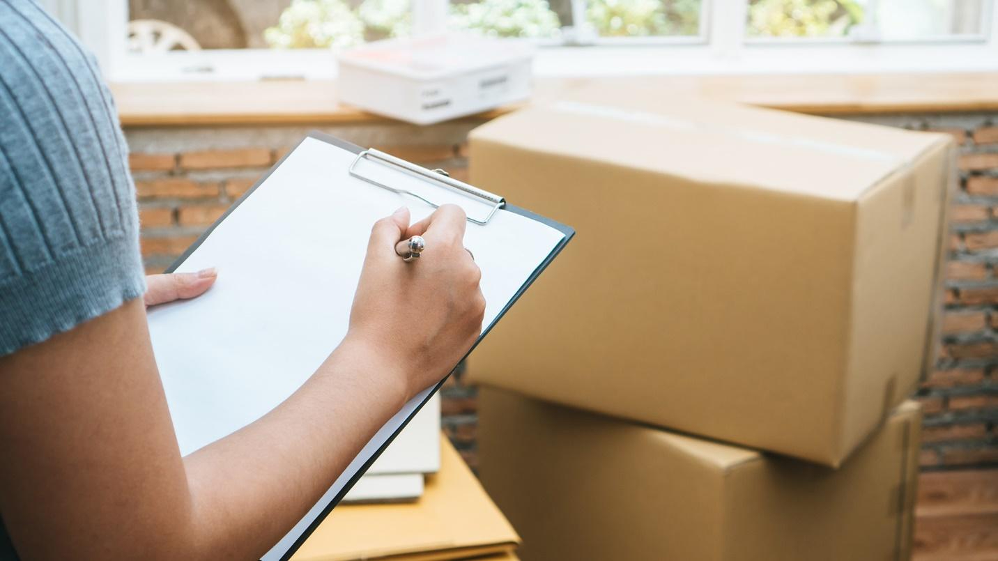 A person writing notes on a clipboard with boxes, a brick wall, and a window in the background.
