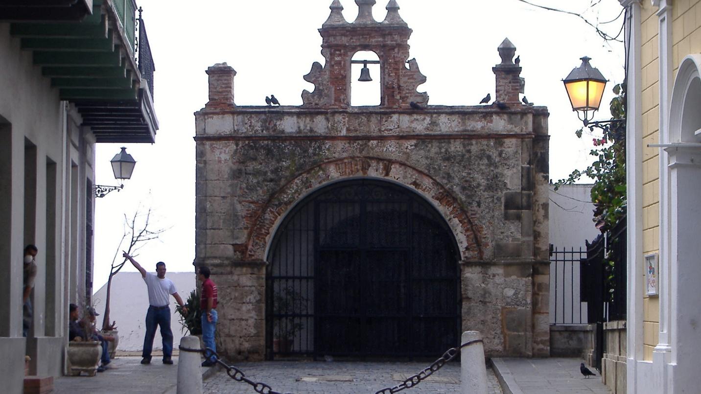 Two men and several pigeons stand by the dark entrance of Capilla del Cristo in a section about the chapel’s features.