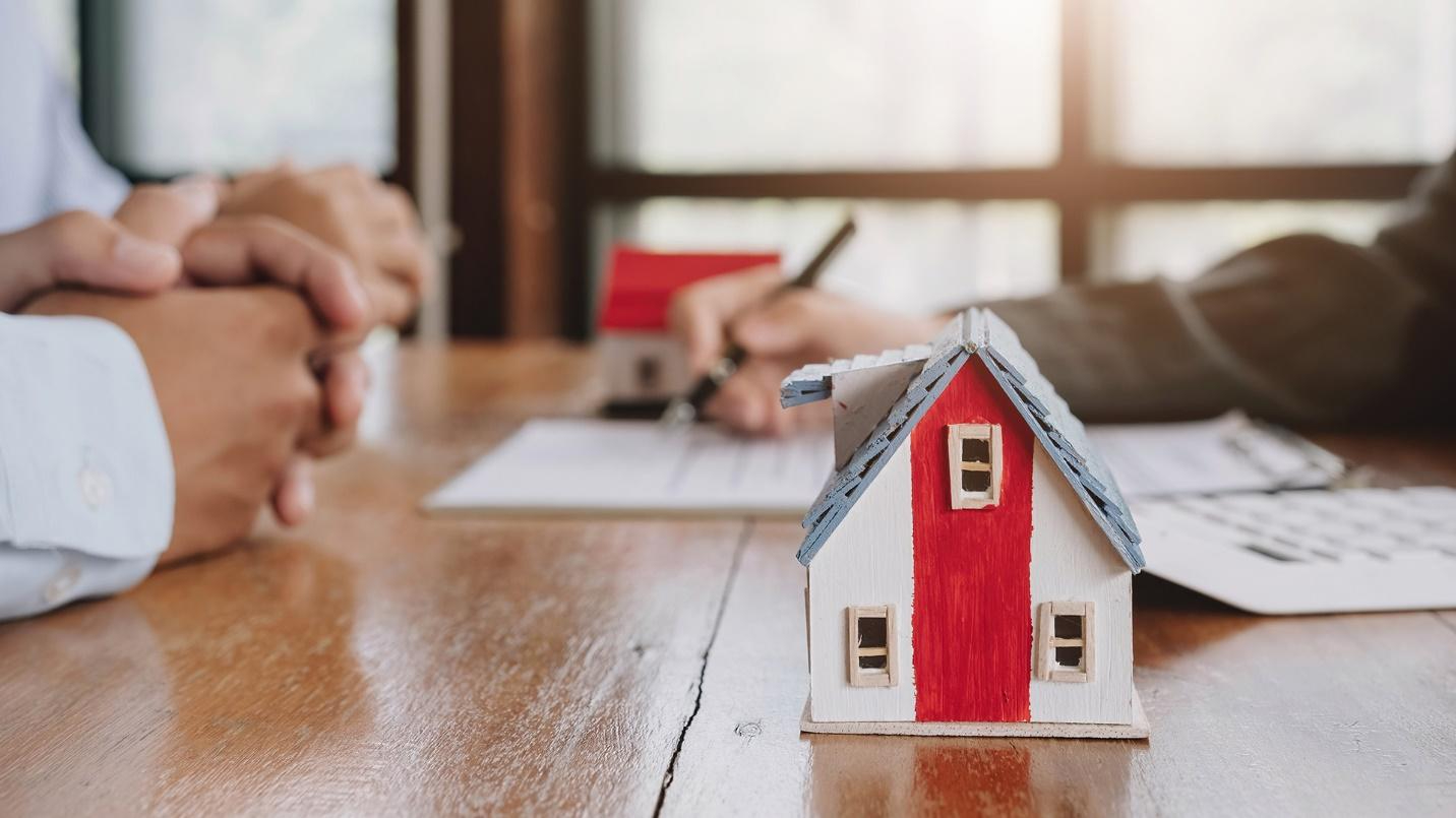 People sitting at a desk while one person writes notes on a pad of paper, with a model of a house in the foreground.