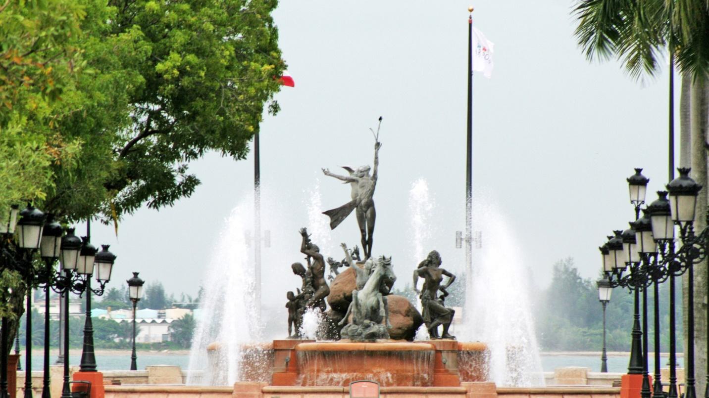 The bronze statues of Raices Fountain by jets of water, trees, and street lamps in a section describing Paseo de la Princesa.