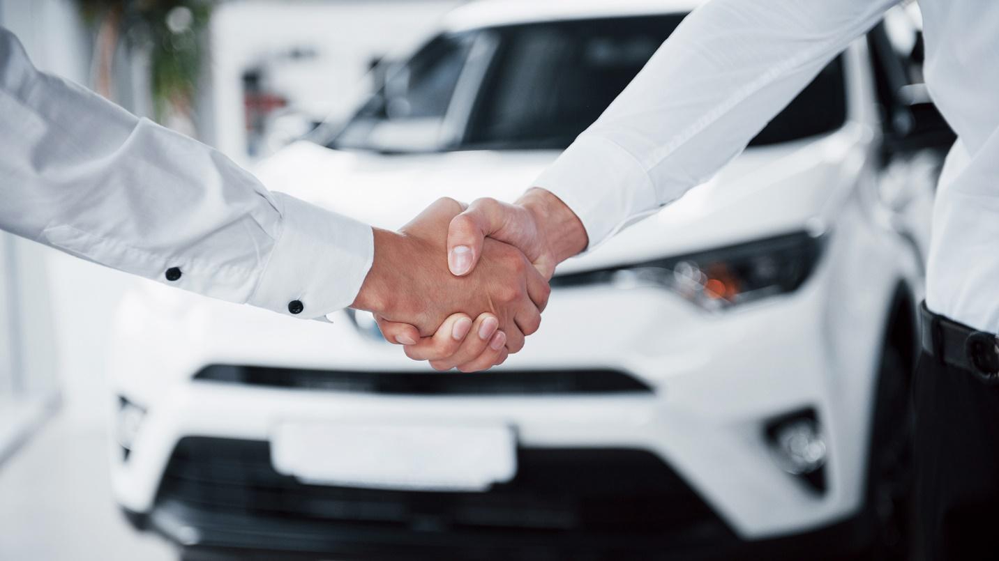 Two people wearing white shirts and shaking hands in the foreground with a white car in the background.
