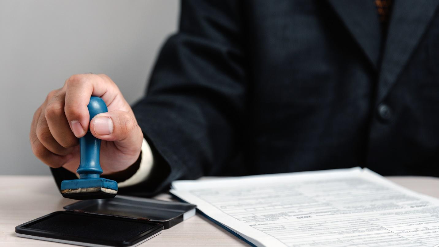 A person wearing a black suit and holding a stamp above an ink pad at a desk with documents.