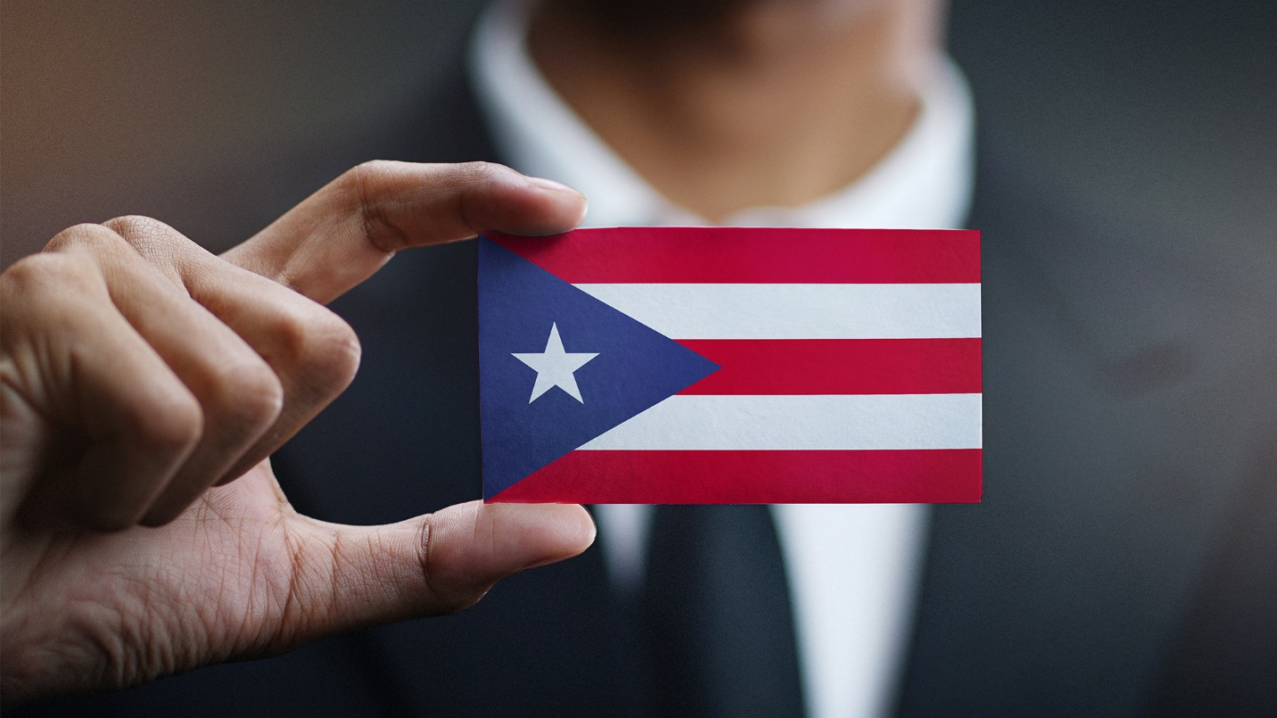 A man in a suit holding a card that depicts a small flag of Puerto Rico.