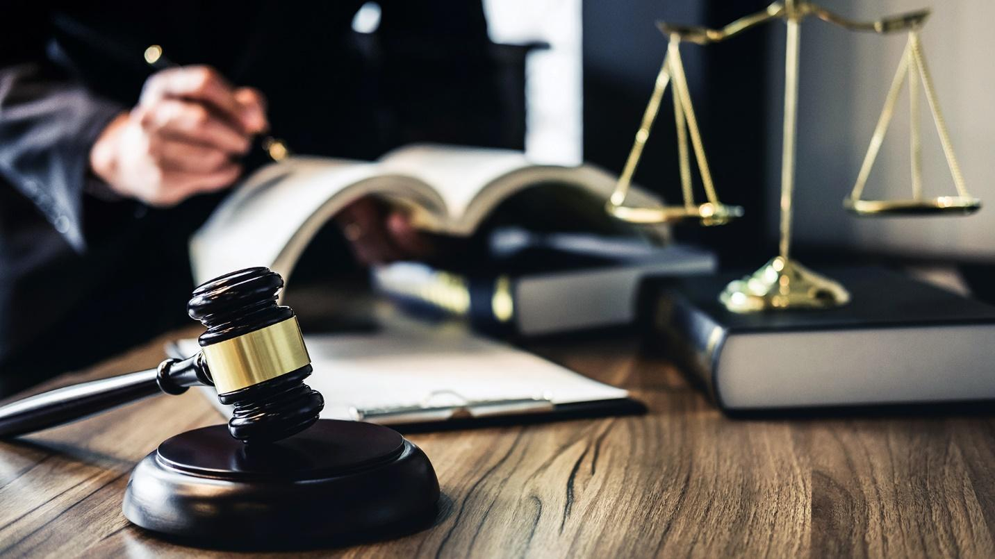A person wearing black and holding a pen and a book at a desk with scales, a gavel, books, and a clipboard.