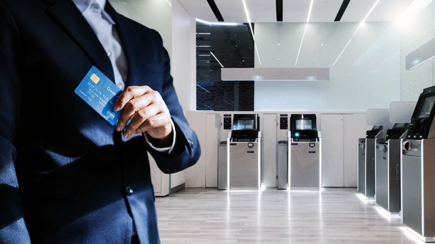 Man in a suit holding a credit card inside a room with self-servicing banking kiosks in the background.