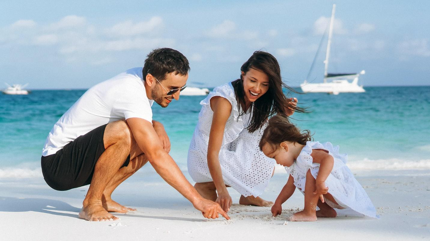 A man, woman, and young girl playing in the sand at the beach, with the sea, sky, and boats in the background.