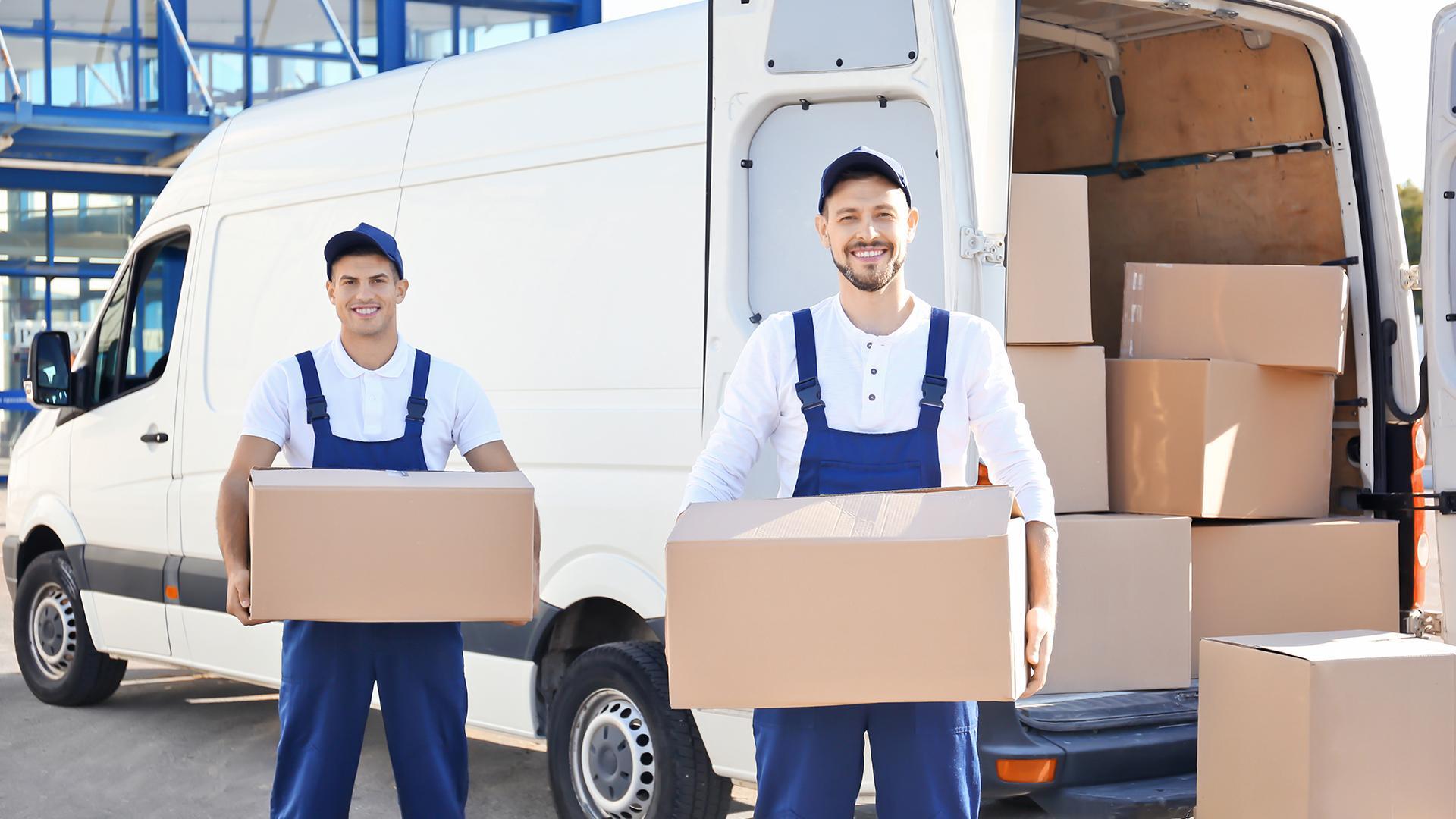 Two smiling movers wearing blue and white carry cardboard boxes behind a white van loaded with more boxes.