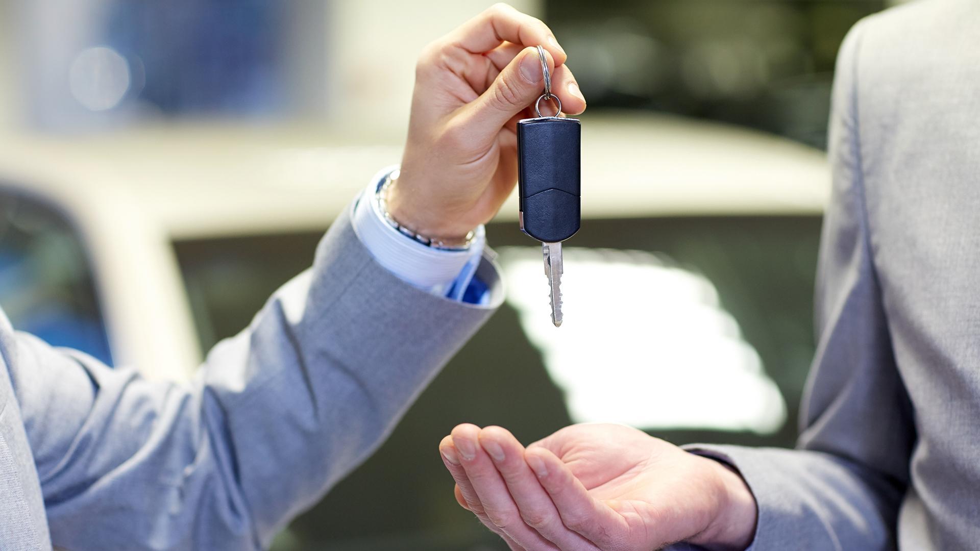 One person wearing a gray suit hands a key to another person wearing a gray suit, with a car in the background.