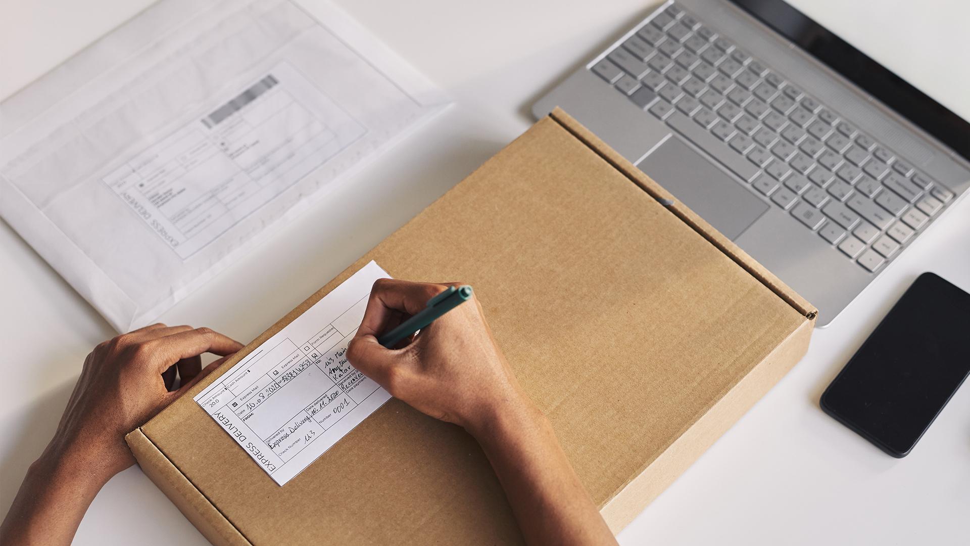 A person filling out a form marked “Express Delivery” on a cardboard box beside a laptop, smartphone, and mailing envelope.