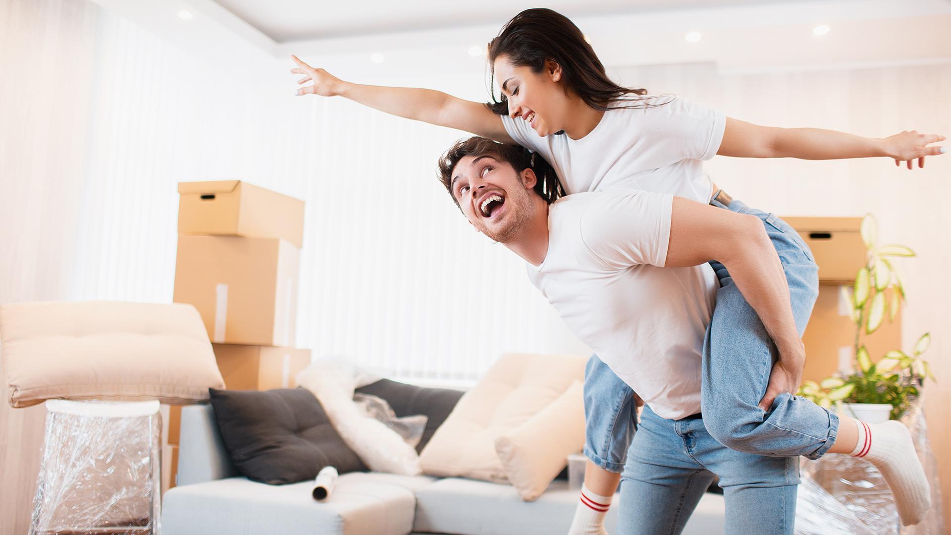 A young couple wearing white shirts and blue jeans celebrate their move in a room that contains stacks of boxes, a couch, and pillows.