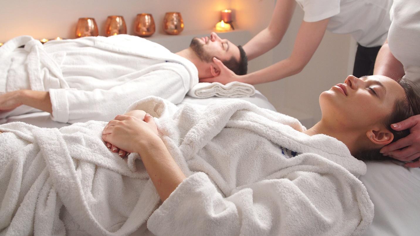 A man and woman dressed in white robes lie flat on their backs with their eyes closed as their heads are massaged.

