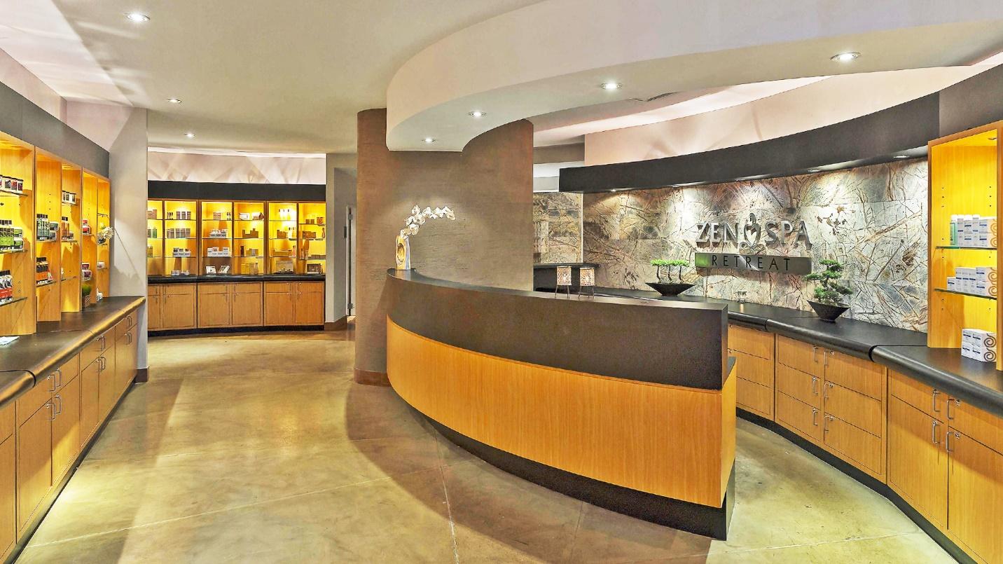 The lobby of Zen Spa Retreat, with a curved wall desk, marble floor, and walls with rows of spa products on display above wood storage cabinets.
