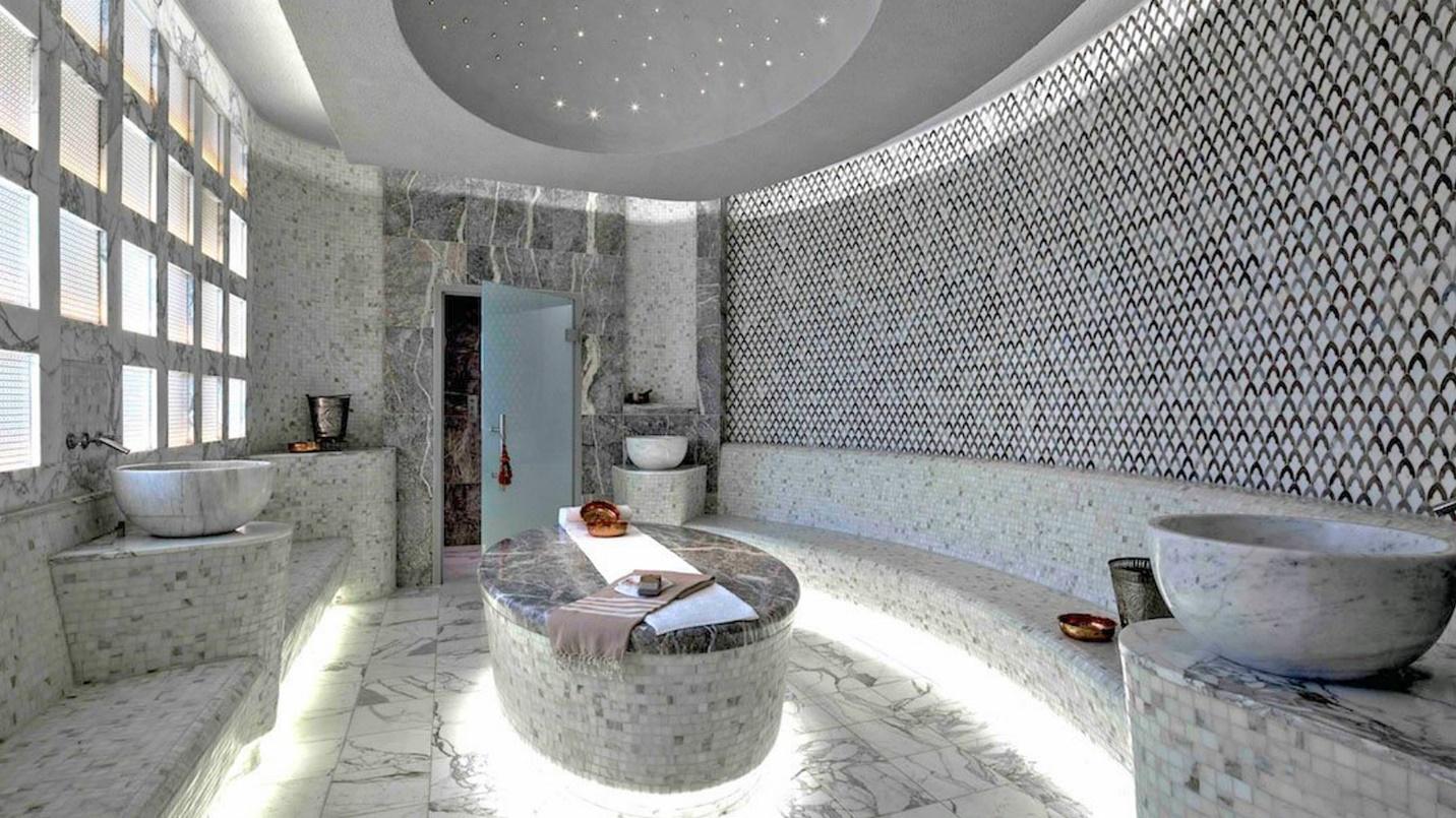 A room with floor-to-ceiling gray tiled surfaces, a curved wall and bench, vessel sinks, and indirect floor and ceiling lighting.
