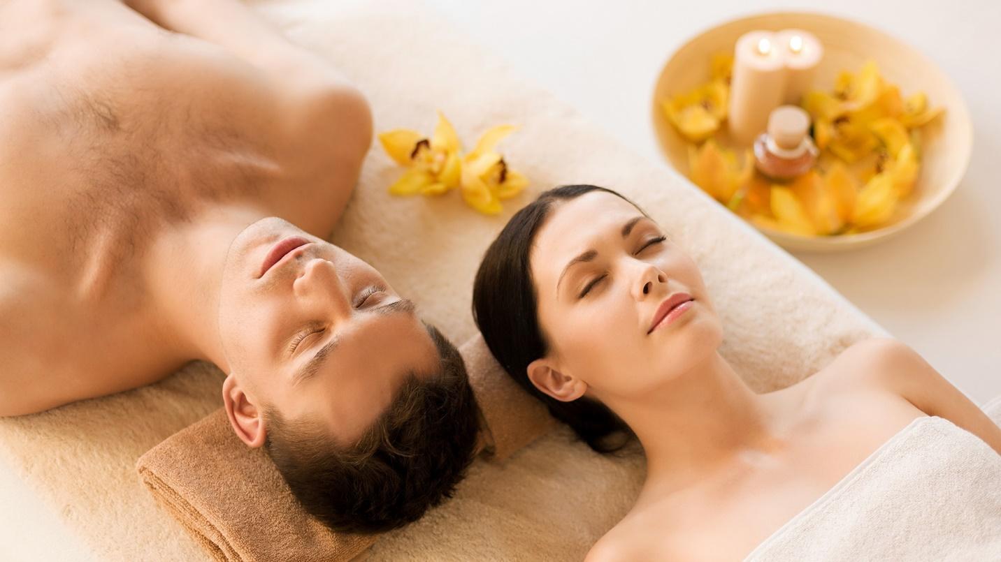A man and woman with eyes closed rest their heads on towels, with flower petals and candles in the background.
