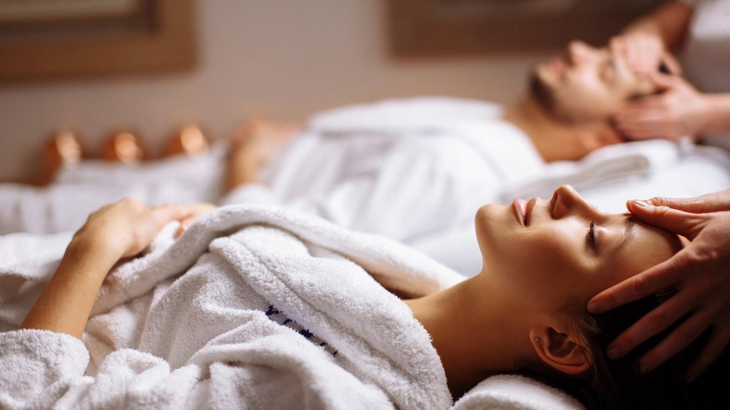 A man and woman dressed in white robes lie flat on white beds as their heads are massaged.
