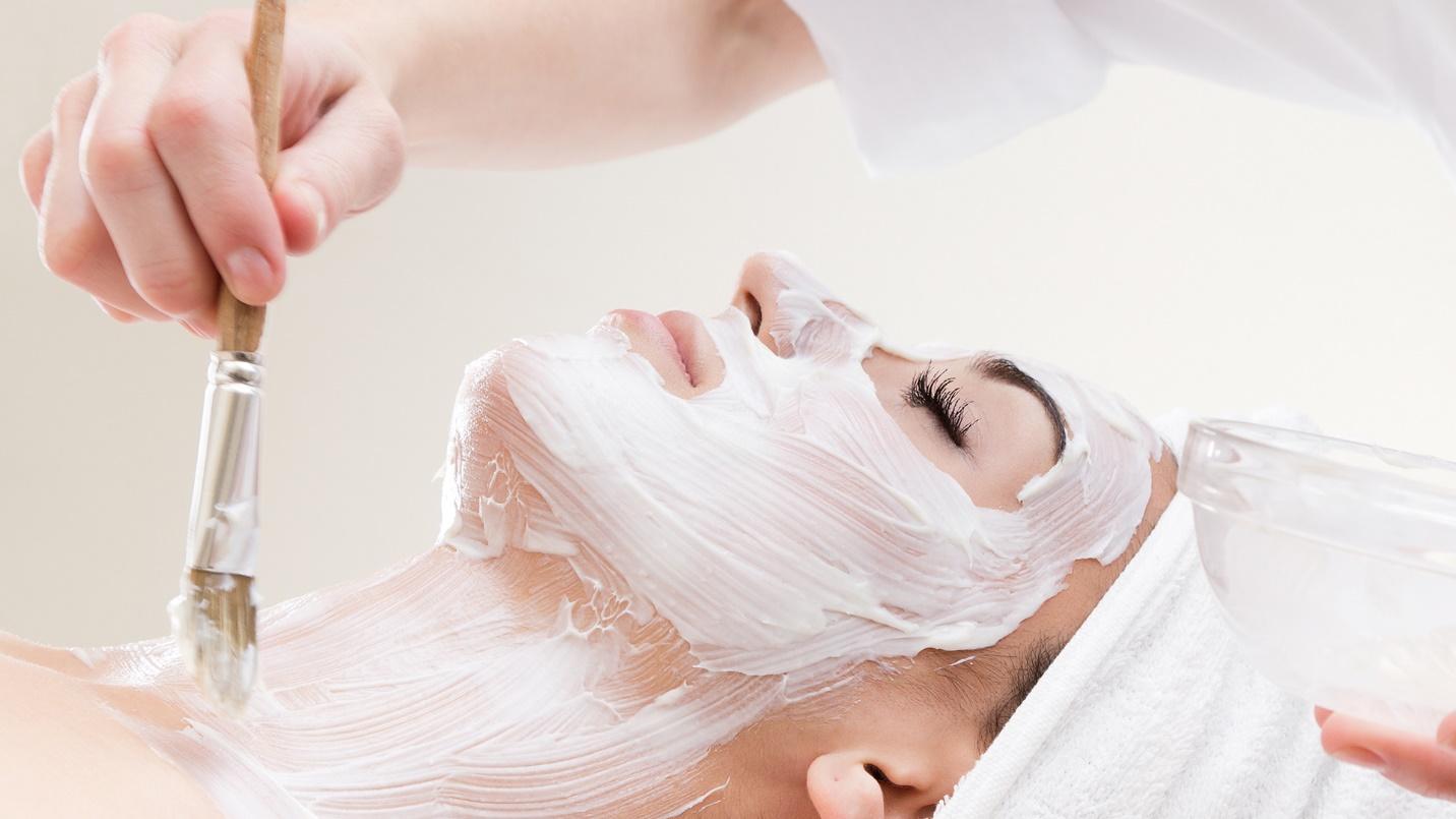 A hand using a brush to apply white lotion to the face and neck of a reclining woman.
