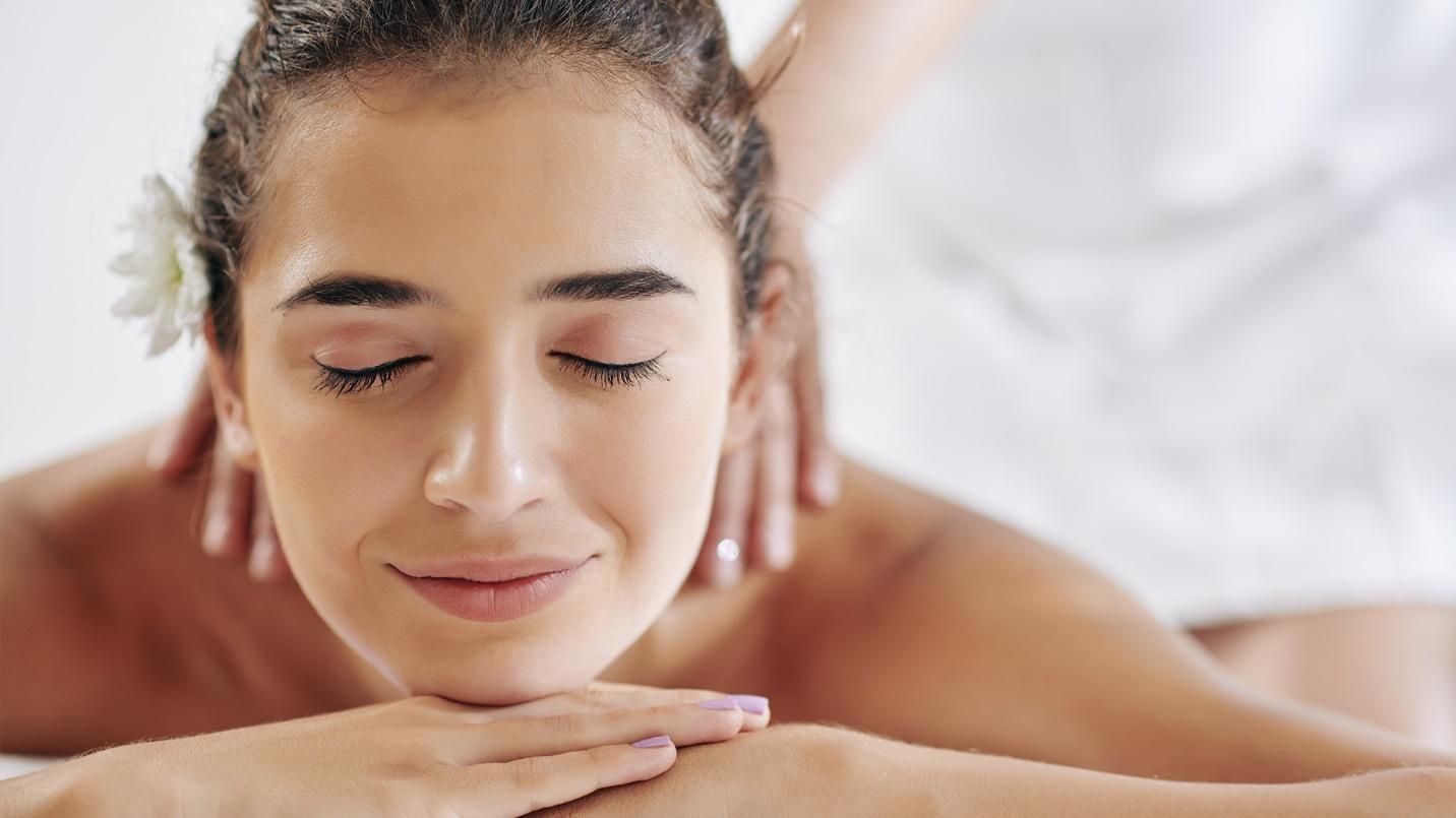 A smiling brunette woman with closed eyes lies flat on her stomach as hands massage her neck.
