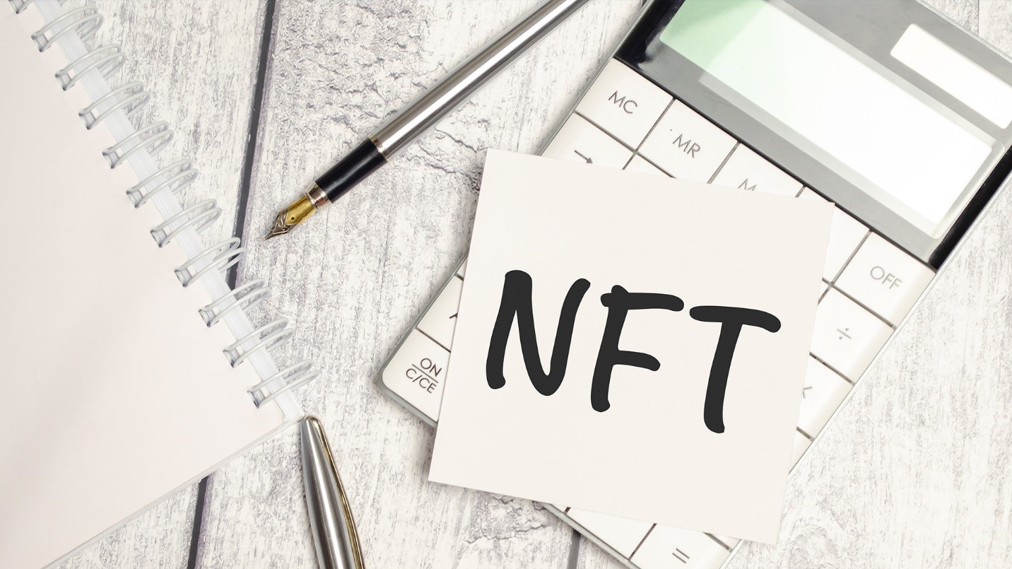 A white wooden plank table holds a calculator, pens, a notebook, and a small piece of paper that shows the letters “NFT,” in a section describing how and when NFT transactions are taxed.