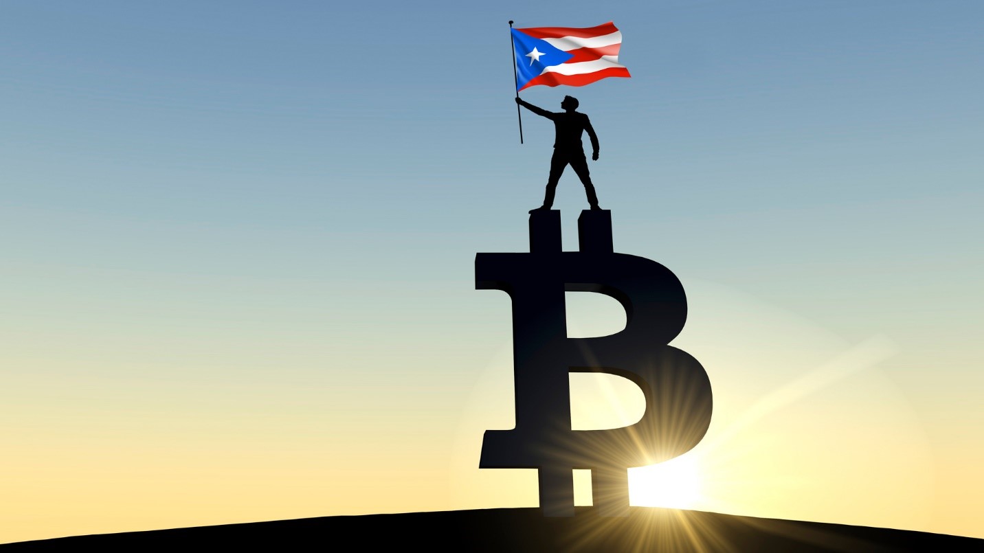 A man holding a Puerto Rican flag while standing on top of a giant Bitcoin symbol in a section that says Puerto Rico is a great place to live for crypto investors.