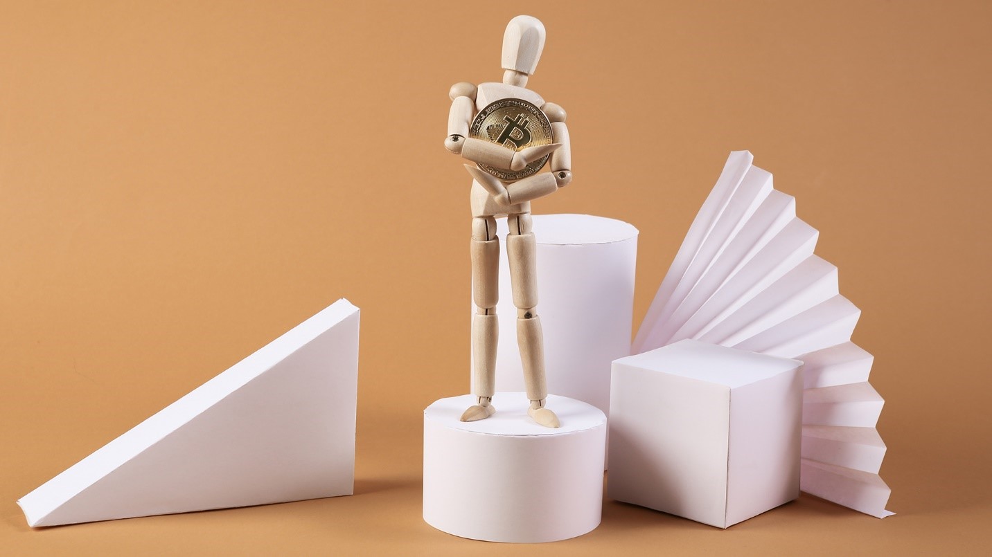 A wooden figure clutches a golden Bitcoin and is surrounded by white 3D geometric figures in a section describing the growing popularity of NFTs.
