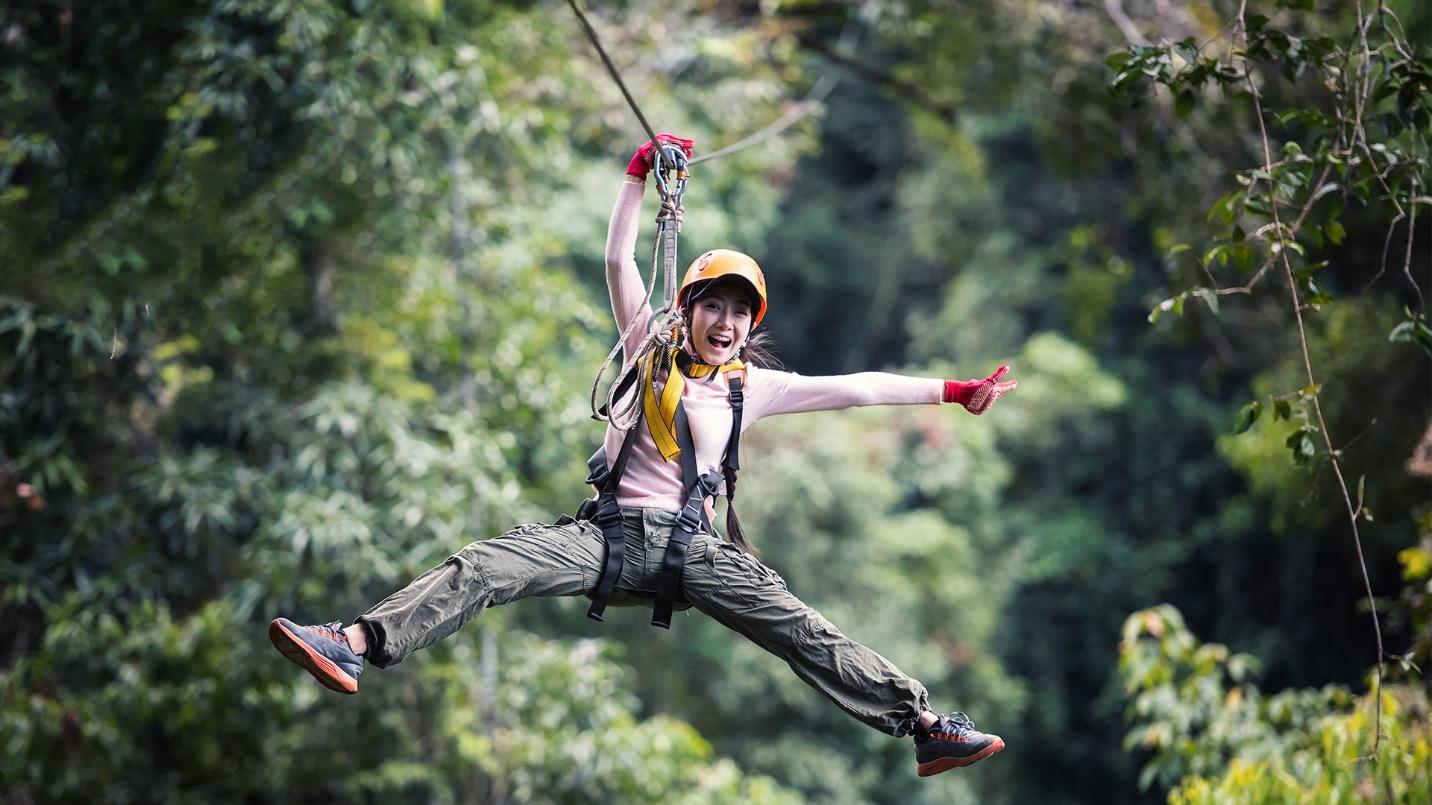 An excited young woman wearing a helmet, gloves, and safety harness zip lines through the trees in a section describing other zip line tour options.