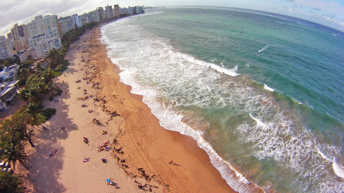Blue-green waves lap at sandy shores framed by beachgoers, high-rise buildings, and trees at Ocean Park Beach in Puerto Rico.
