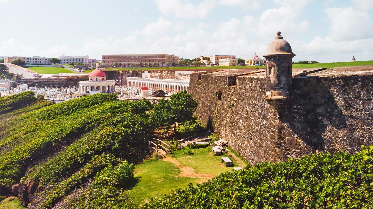 The limestone walls of a Spanish fort surrounded by vegetation and a cemetery at San Juan National Historic Site.