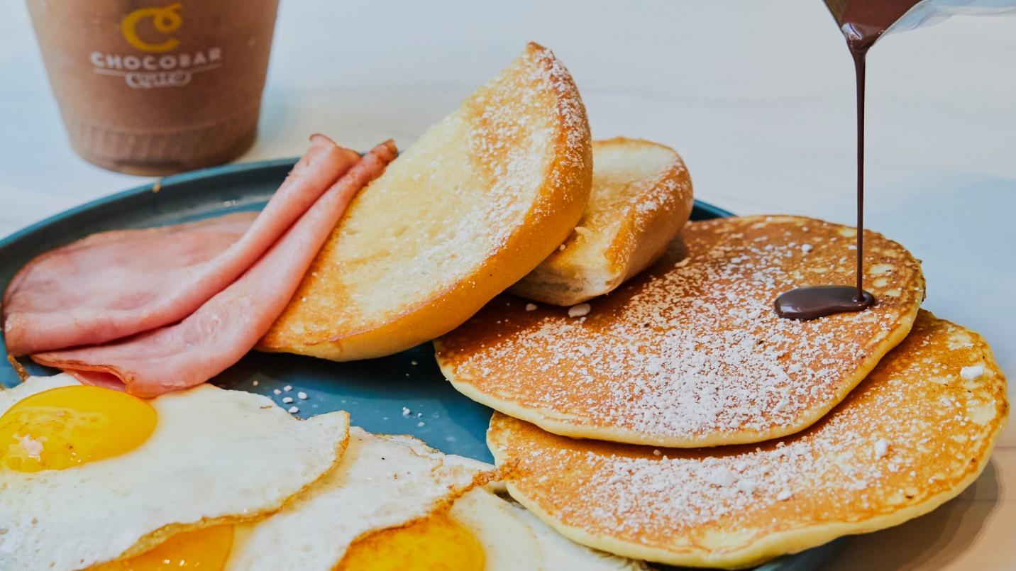 Chocolate syrup is drizzled on pancakes next to ham, fried eggs, and fried bread in a section describing the chocolate-focused Chocobar Cortes restaurant.