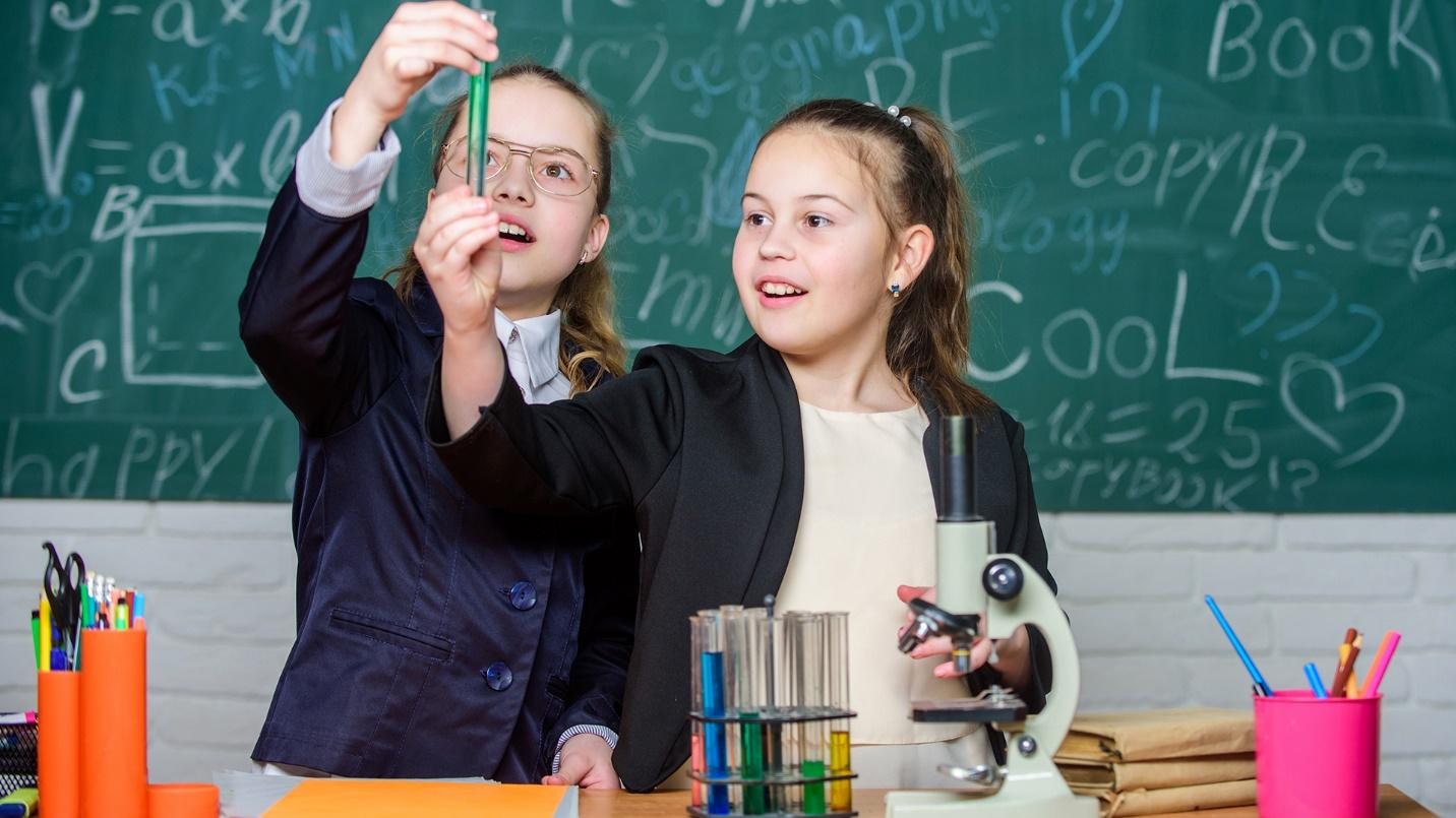 Two young female students learning in a classroom next to a microscope, school supplies, and chalkboard.