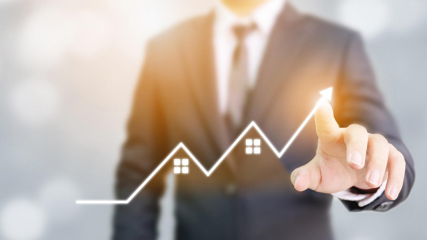 A man in a suit pointing at an image of houses beneath a line graph pointing upward.