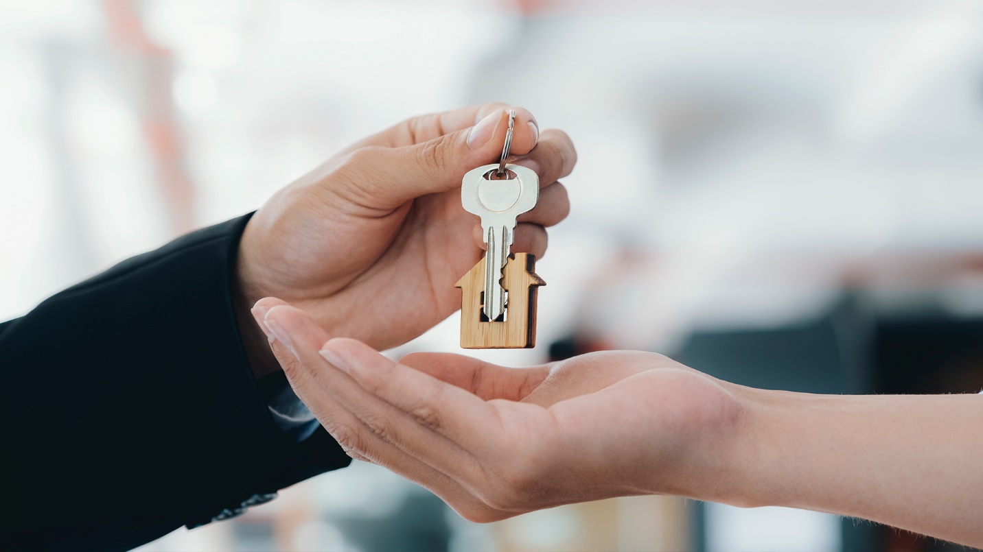 A person drops a key with a house-shaped keychain into another person’s outstretched hand.