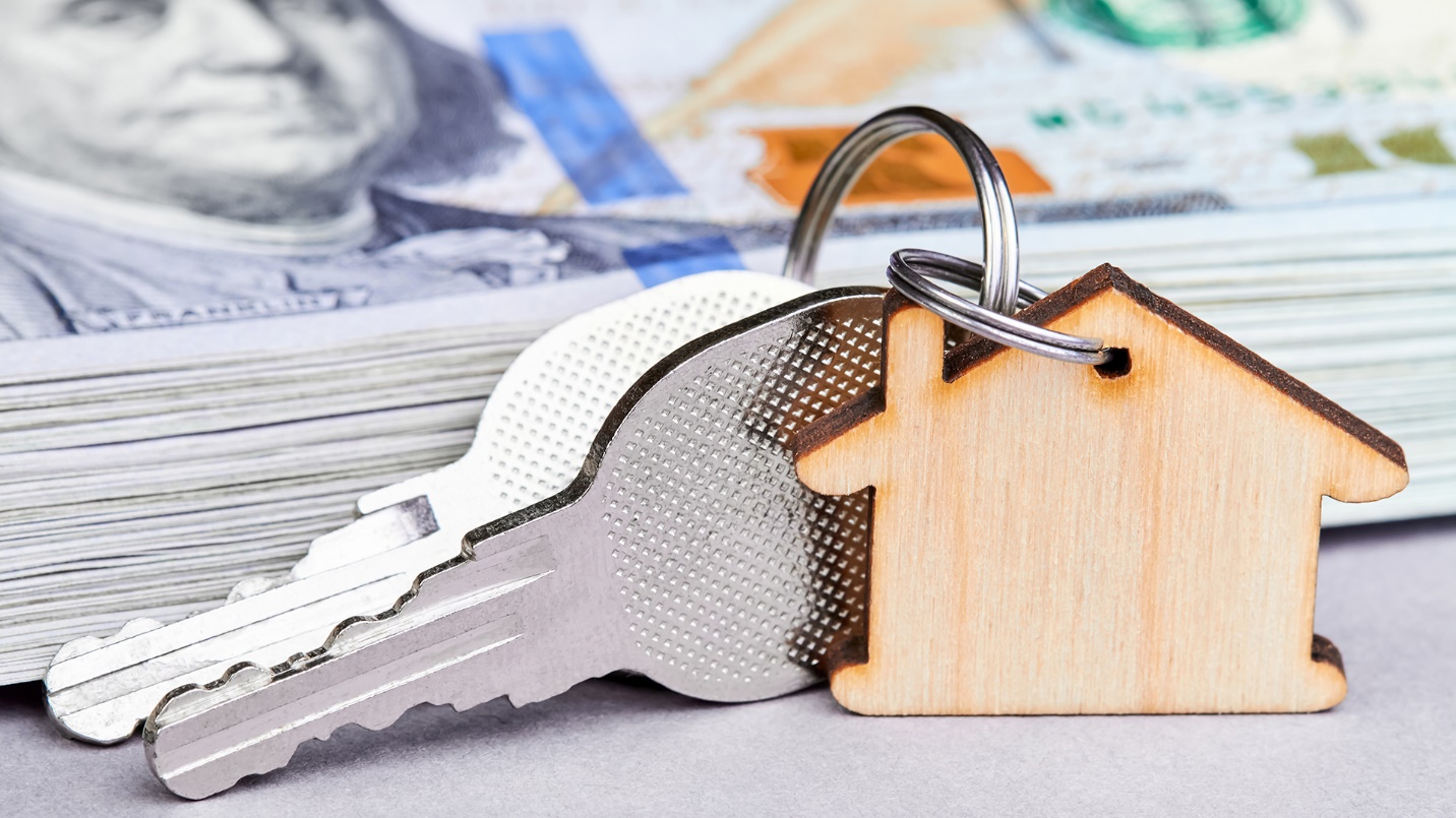 Silver keys on a house-shaped keychain beside a stack of one-hundred-dollar bills.