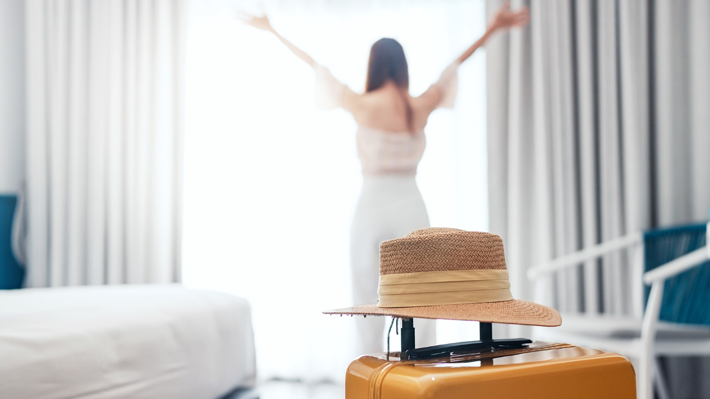 In the foreground, a hat rests on a suitcase, and in the background, a woman with outstretched arms stands by a window, bed, and chair.
