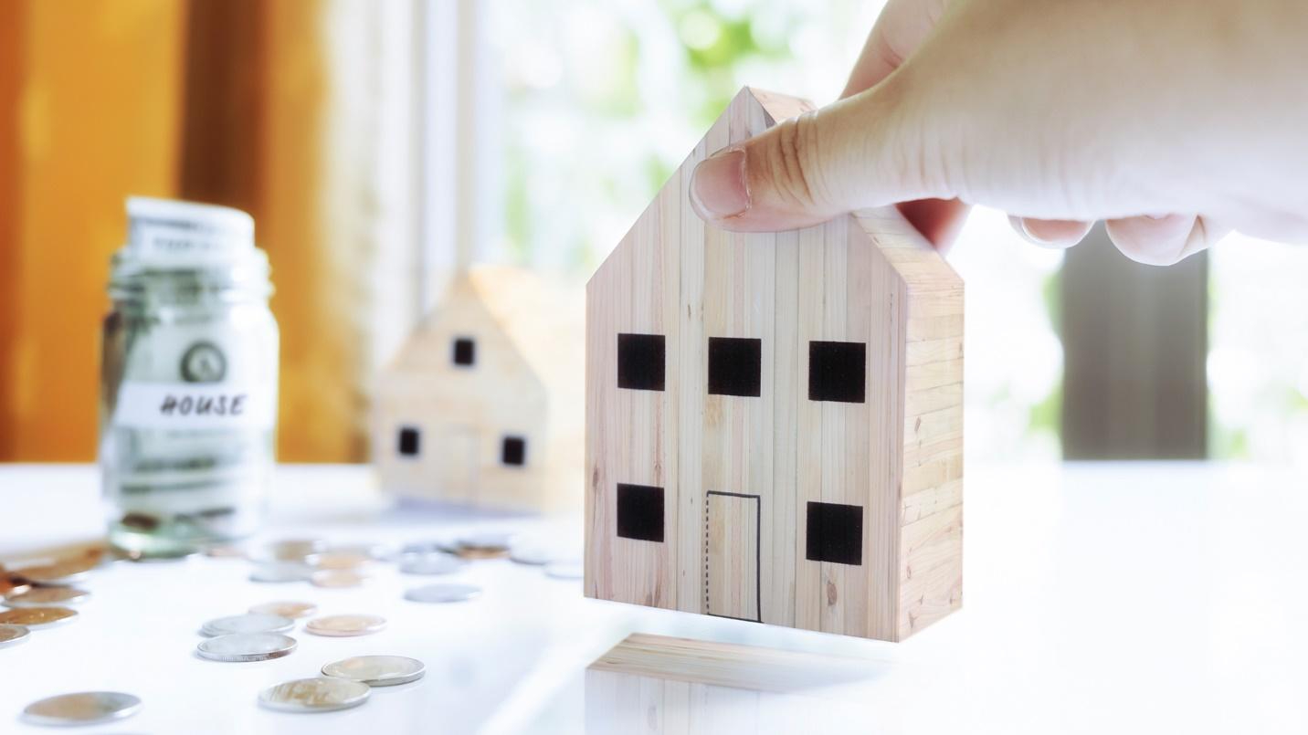 Wooden models of houses on a table beside coins and a jar labelled “House” that contains cash.
