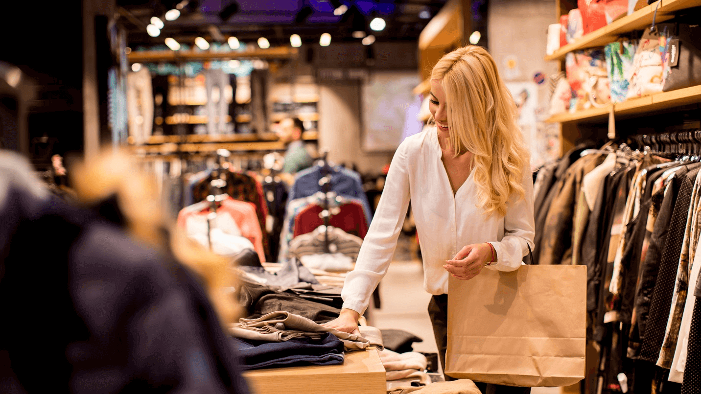 A blonde woman dressed in white holds a brown shopping bag while browsing in a clothing store.