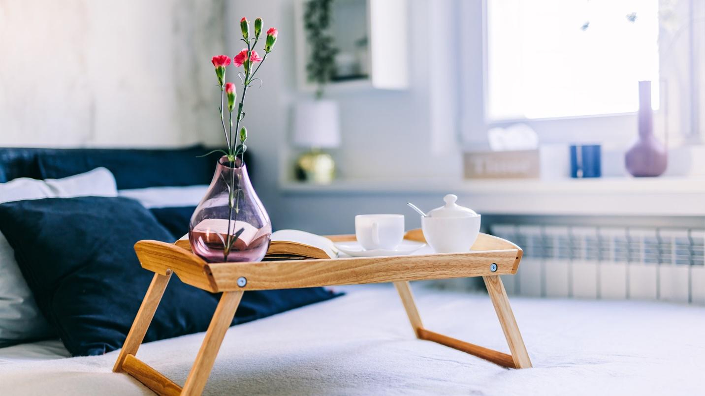 A tray holding a cup and saucer, container of sugar, an open book, and a vase resting on top of a bed.
