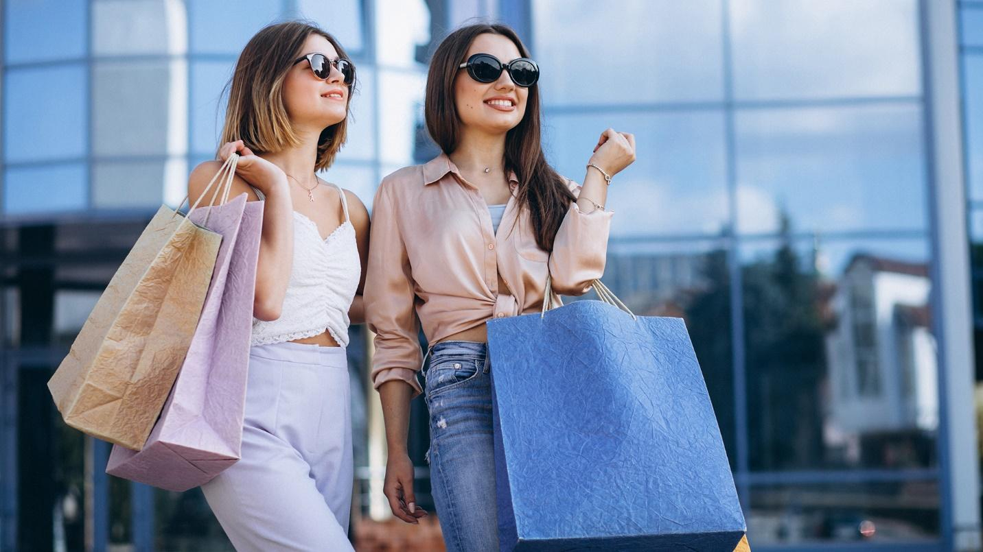 Two women wearing sunglasses and holding shopping bags outside.