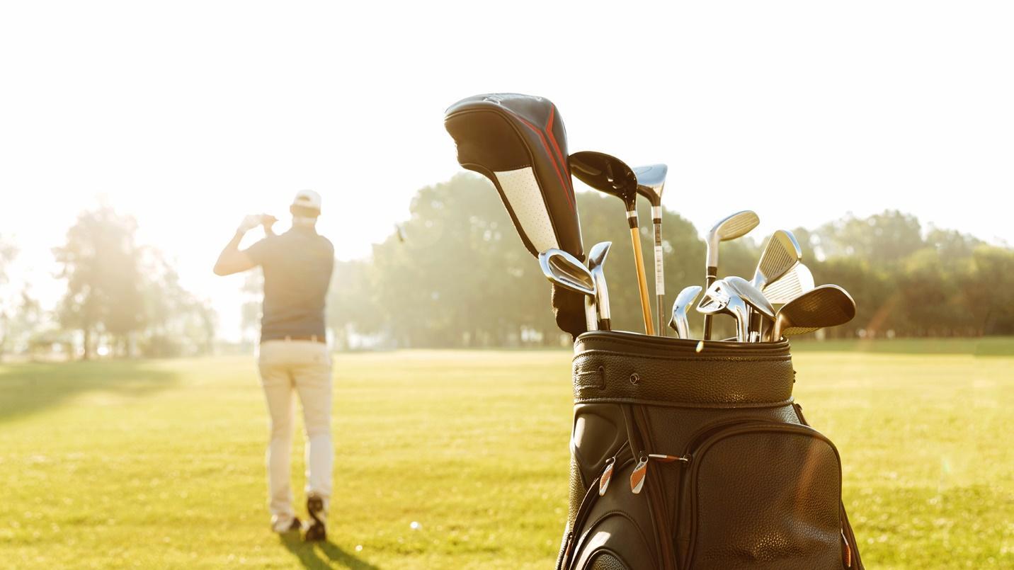 A close-up of a set of golf clubs at a course with a man taking a swing in the background in a section about the area’s PGA courses, outdoor activities, and natural beauty.
