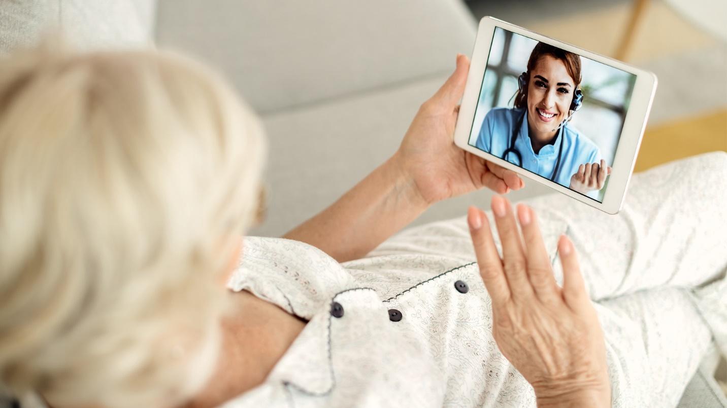 An elderly woman sitting on a couch interacts with a female healthcare worker via her tablet in a section about health care.