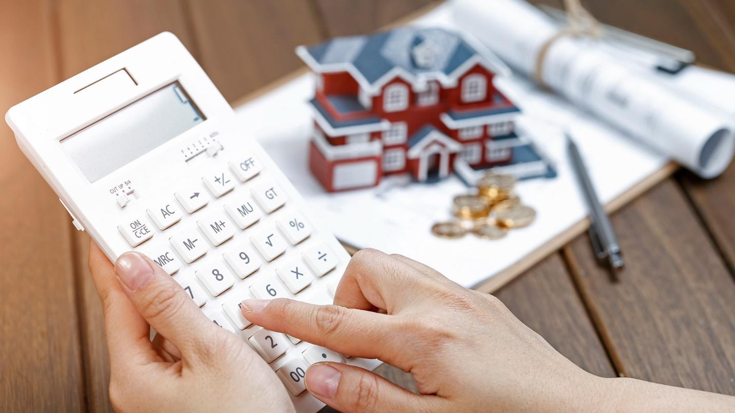 A person holds a calculator with a model house, clipboard, and pen in the background in a section about real estate prices.