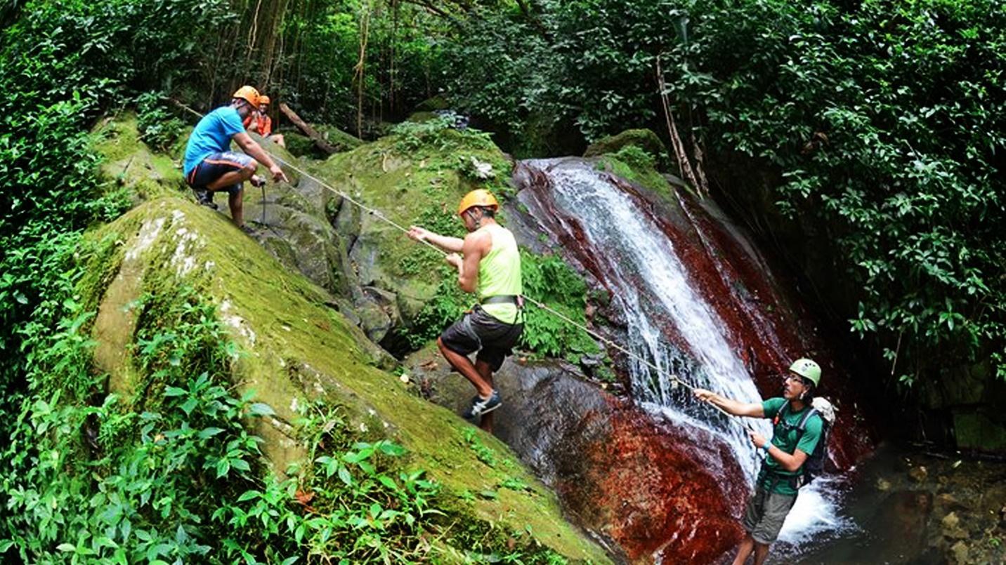 Four adventurers wearing helmets use ropes to scale large mossy rocks beside a waterfall in a section about Rocaliza Adventure Tours. 